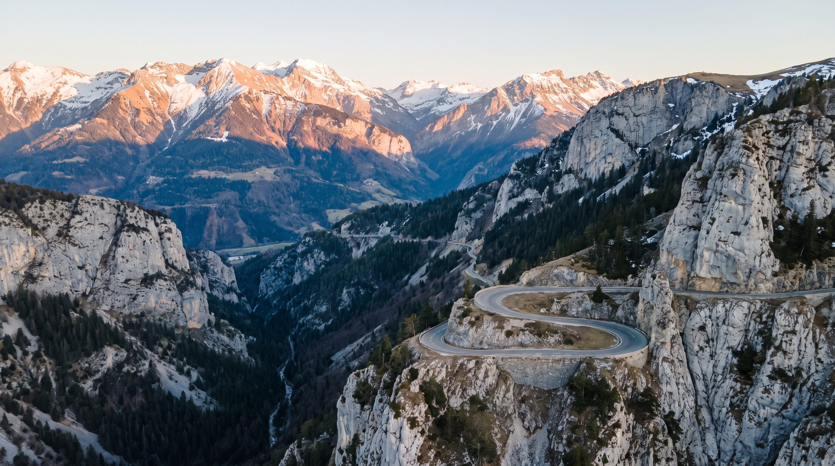 A sweeping alpine road carved into a limestone hillside with snow capped peaks beyond