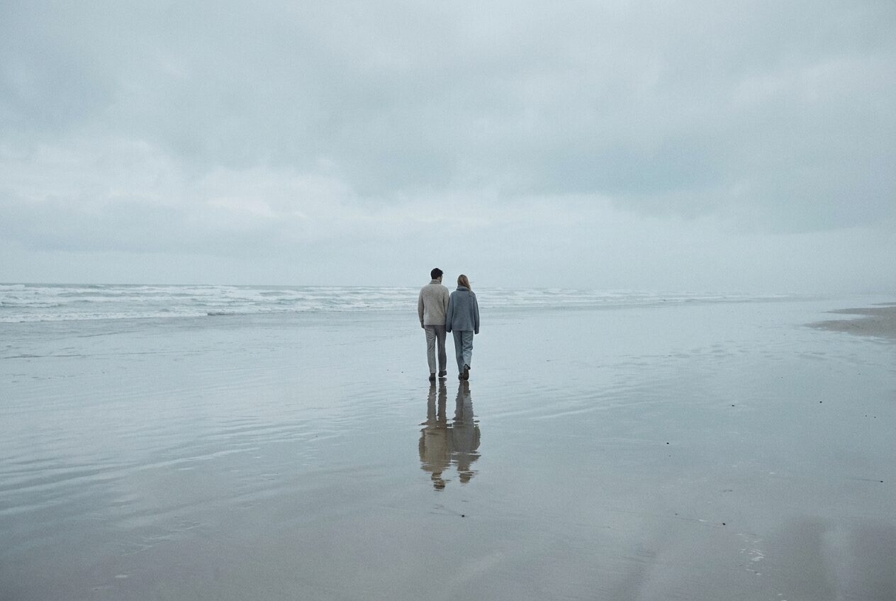 Two models in oatmeal and slate knitwear walking a wide empty tideline